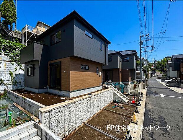 【現地外観写真】黒と木目調のコントラストが美しいモダンな新築戸建て。道路より一段高い敷地にあり、陽当たりや風通しも良好です。青空に映えるスタイリッシュな邸宅で、心豊かな新生活を始めませんか。