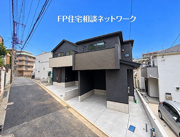 【前面道路含む現地写真】黒の外壁と木目調のバルコニーが印象的な戸建て住宅です。ビルトインガレージを備え、天候を問わず車の乗り降りが可能です。