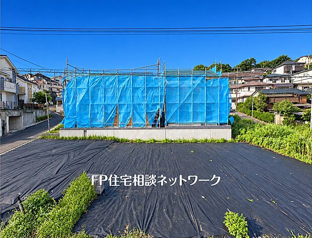【前面道路含む現地写真】角地に位置する建築中の建物の様子です。周囲は足場と養生シートで覆われ、完成に向けて工事が進められています。接道する道路の状況や隣地との高低差など、周辺の環境が確認できます。