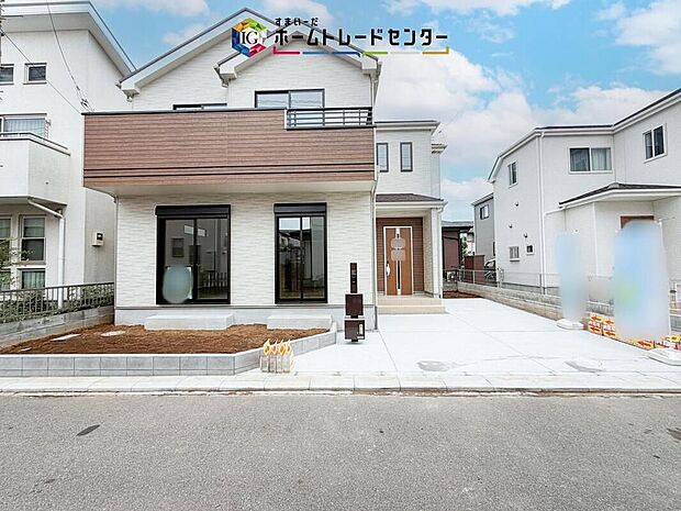 【現地外観写真】白を基調に木目調のバルコニーが映える外観。建物正面と駐車スペースを一度に確認でき、玄関までの動線や住まい全体の雰囲気もイメージしやすい一枚です。正面の大きな窓もよく印象的に映ります。