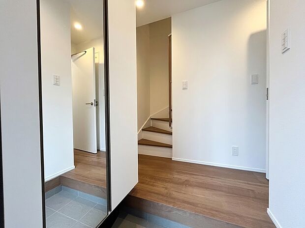 【玄関】お出かけ前の全身チェックができる大型姿見付きの玄関!温かみのある木目調のフローリングが心地よく家族を迎えます。奥行きを感じられる開放的な設計で、2階へとスムーズにつながる階段が配された機能的な動線です