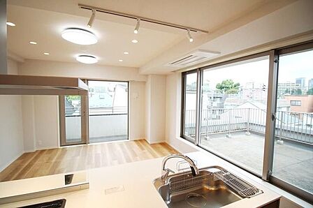 手前にシンクを備えた対面式キッチンが配置されており、天井にはビルトインエアコンやライティングレール、ダウンライトが設置されています。大きな窓や勝手口風の扉からはバルコニーへ出られる造りです。