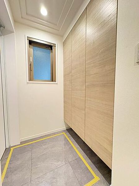 グレー調のタイルが敷かれた玄関には、壁面いっぱいに木目調の大型シューズボックスが備え付けられており、すりガラスの小窓や天井のダウンライトが設置された落ち着いた内装デザインとなっています。