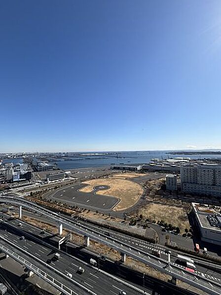 屋外を撮影した景色で、手前には幹線道路や高架橋、中央には広場や建物が配置されており、その奥には海や大きな橋を含んだ沿海部の風景が水平線まで広がっています。