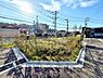 外観：遮るものが少なく開放感のある現地土地写真です。周囲に植栽が施されており緑が感じられます。道路に面しており角地の形状が特徴的です。日当たりも良好な環境です。