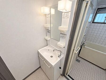 洗面台は壁付けタイプで空間を有効に活用できます。鏡の両側に照明があり明るさが確保されています。収納スペースが備えられ小物整理に便利です。浴室と洗面所が隣接していて動線もスムーズです。