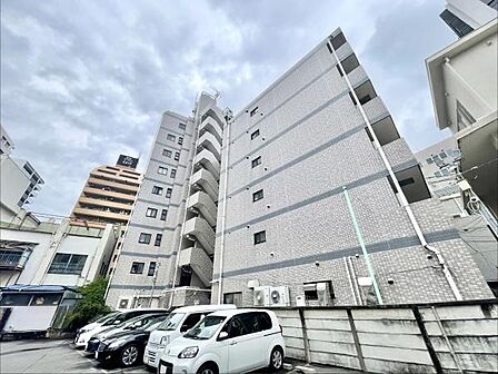 シンプルなデザインの外観が特徴的です。敷地内には複数台分の駐車スペースがあります。周囲には建物が立ち並び都市的な雰囲気を感じられます。建物の階数が多く存在感があります。清潔感のある外壁が印象的です。