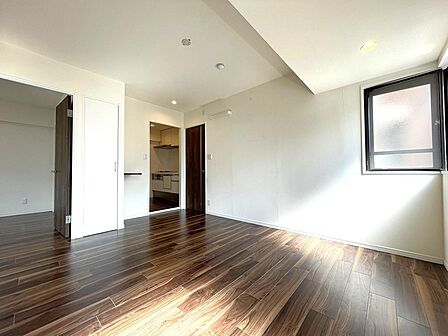 深みのあるシックな木目調フロアと白のクロスの対比が美しいリビング。窓から柔らかな光が差し込み、リビング収納やキッチンへのスムーズな動線も備えた、快適で上質な居心地のよい空間です。