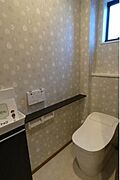 窓が設置されており明るさと換気に配慮されたトイレです。壁紙のデザインが空間にやわらかな印象を与えています。使いやすい位置にペーパーホルダーや棚が設置されています。清潔感を感じる内装です。