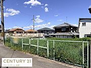 明るい青空の下周囲には住宅が立ち並ぶ落ち着いた現地土地写真です。道路に面しているため車の出入りもスムーズに行えます。開放感のあるフェンスが土地の境界を示しており管理のしやすさがうかがえます。