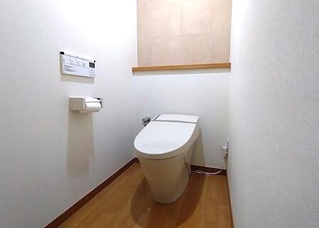 シンプルで清潔感のあるトイレ空間。壁や床の色合いが落ち着いた雰囲気を演出しています。備え付けの棚がアクセントとなっています。操作パネルも見やすい位置に設置されており、無駄のないデザインが特徴です。